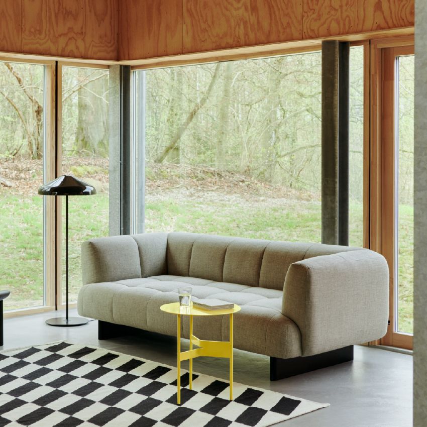 Minimalistische woonkamer met lichtgrijze HAY Quilton bank op zwart onderstel, een geel bijzettafeltje met glas water en boek, zwart-wit karpet en grote ramen met uitzicht op het bos.