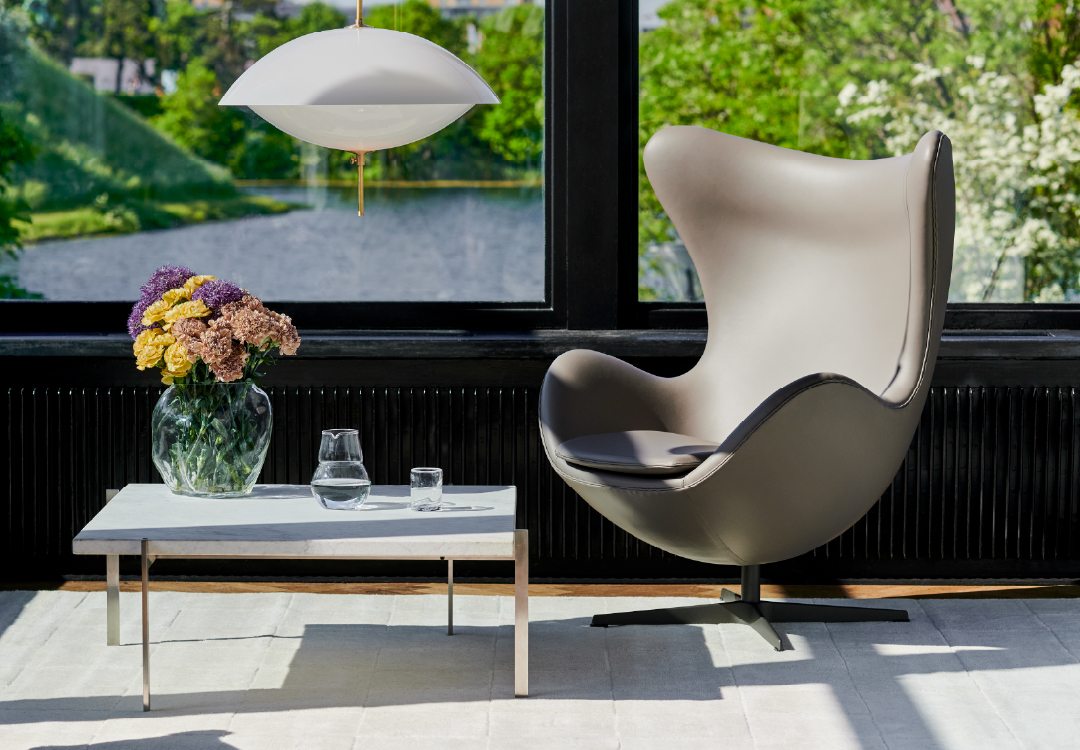 Grijze Egg Chair naast witte salontafel met glazen vaas en kleurrijk boeket, witte hanglamp en uitzicht op water en bomen door groot raam.