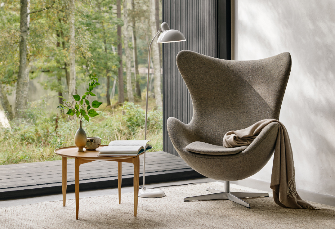 Grijze Egg Chair met beige plaid, houten ronde bijzettafel met boeken en vaas, zilveren booglamp en uitzicht op bos door hoge ramen met zwarte lamellen.
