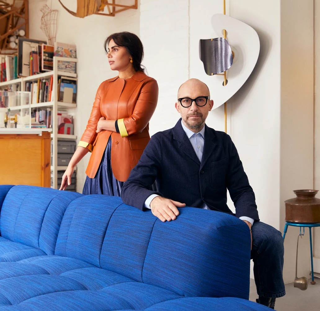 Ontwerpers Doshi Levien bij blauwe designbank Ontwerpers Doshi Levien in een moderne woonstudio, één zittend op een felblauwe geknopte sofa, de ander staand achter de bank bij een boekenkast vol designobjecten