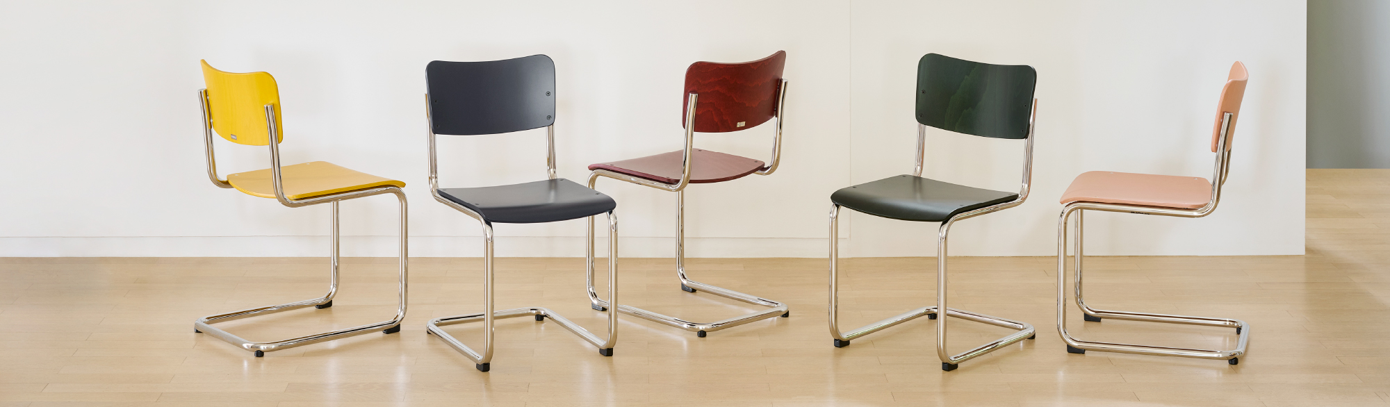 Thonet S43 stoelen in vijf kleuren &ndash; geel, blauw, bordeaux, donkergroen en roze &ndash; met verchroomd buisframe op houten vloer tegen witte wand &ndash; Van Til Interieur.